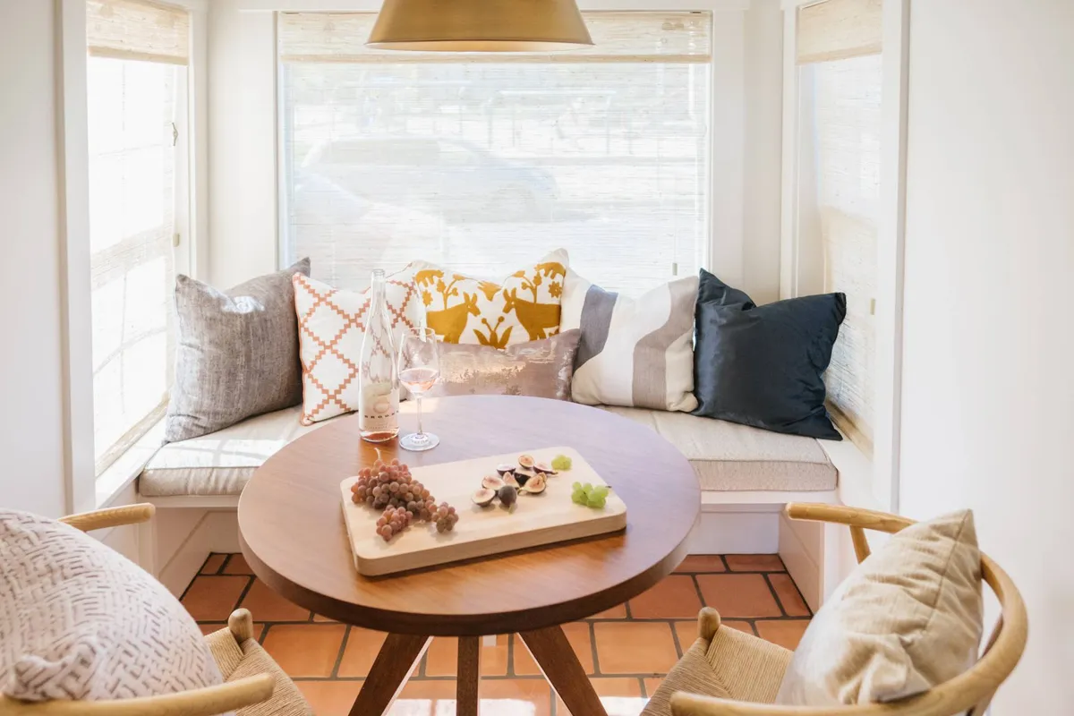 An intimate, light-filled room featuring a round wooden table topped with a platter of fresh fruits, surrounded by comfortable cushions. A large window allows Oregon sunlight to pour in, creating a relaxed ambiance ideal for afternoon tea, casual dining, or quiet reading. The setup adapts well to both social and solo activities.