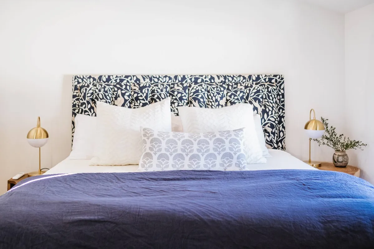 A tranquil bedroom with a decorative headboard, plush white pillows, a textured blue blanket, gold bedside lamps, and a green plant. The bed looks exceptionally comfortable and is staged with high-quality linens and ample pillows, evoking a luxurious retreat.