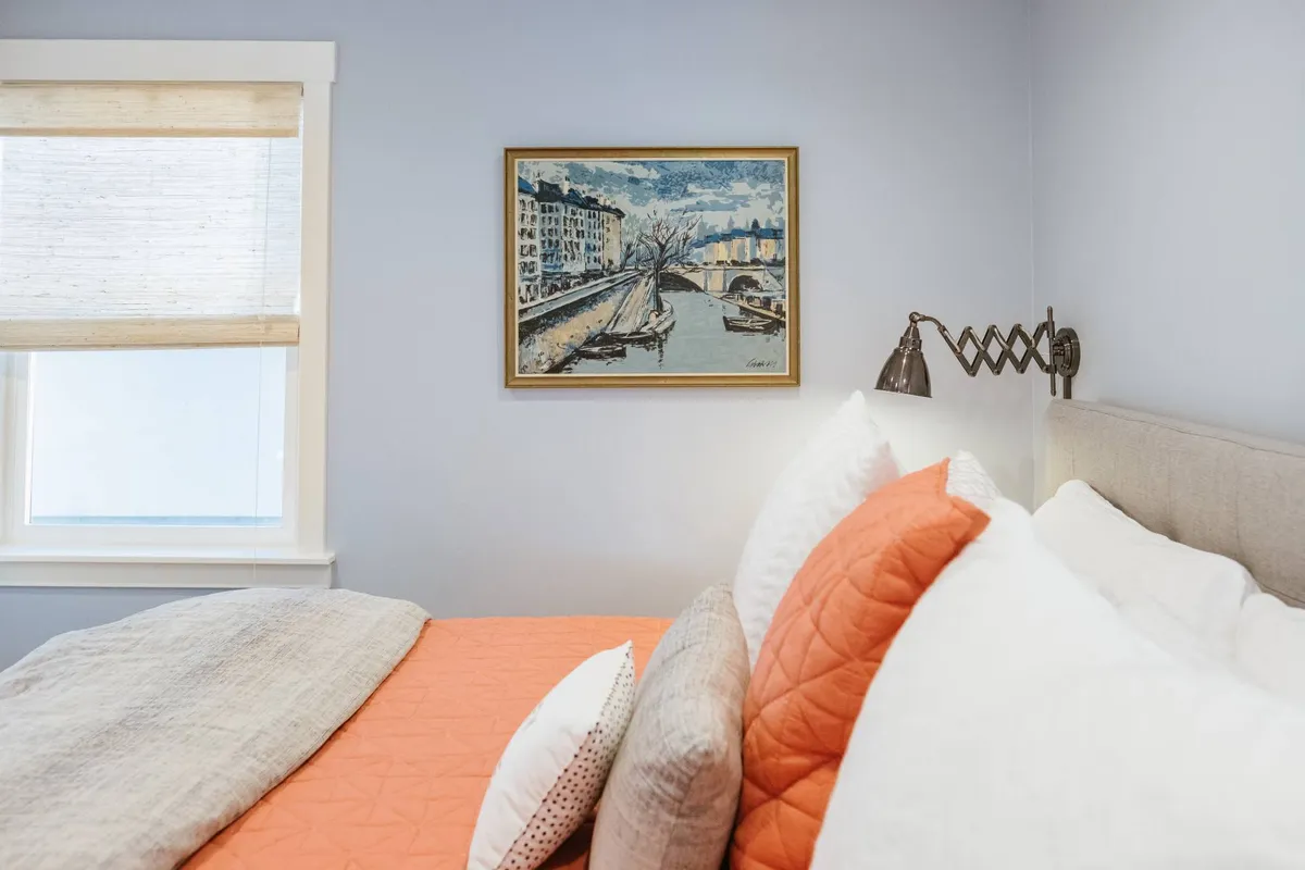 A tranquil bedroom featuring an art piece on the wall, a vibrant orange bedspread, and a cozy bedside light. The ambiance is peaceful, highlighting privacy and personal comfort within the retreat.