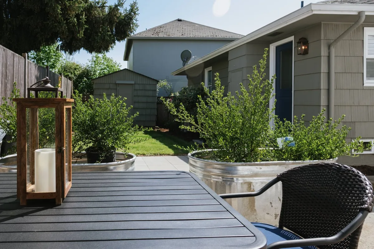 A calm and serene patio scene: a modern black outdoor chair and polished wooden lantern sit among potted plants and silver planters, with the house's exterior in the background. The setup invites relaxation, morning coffee, or an impromptu wine tasting in the garden setting.