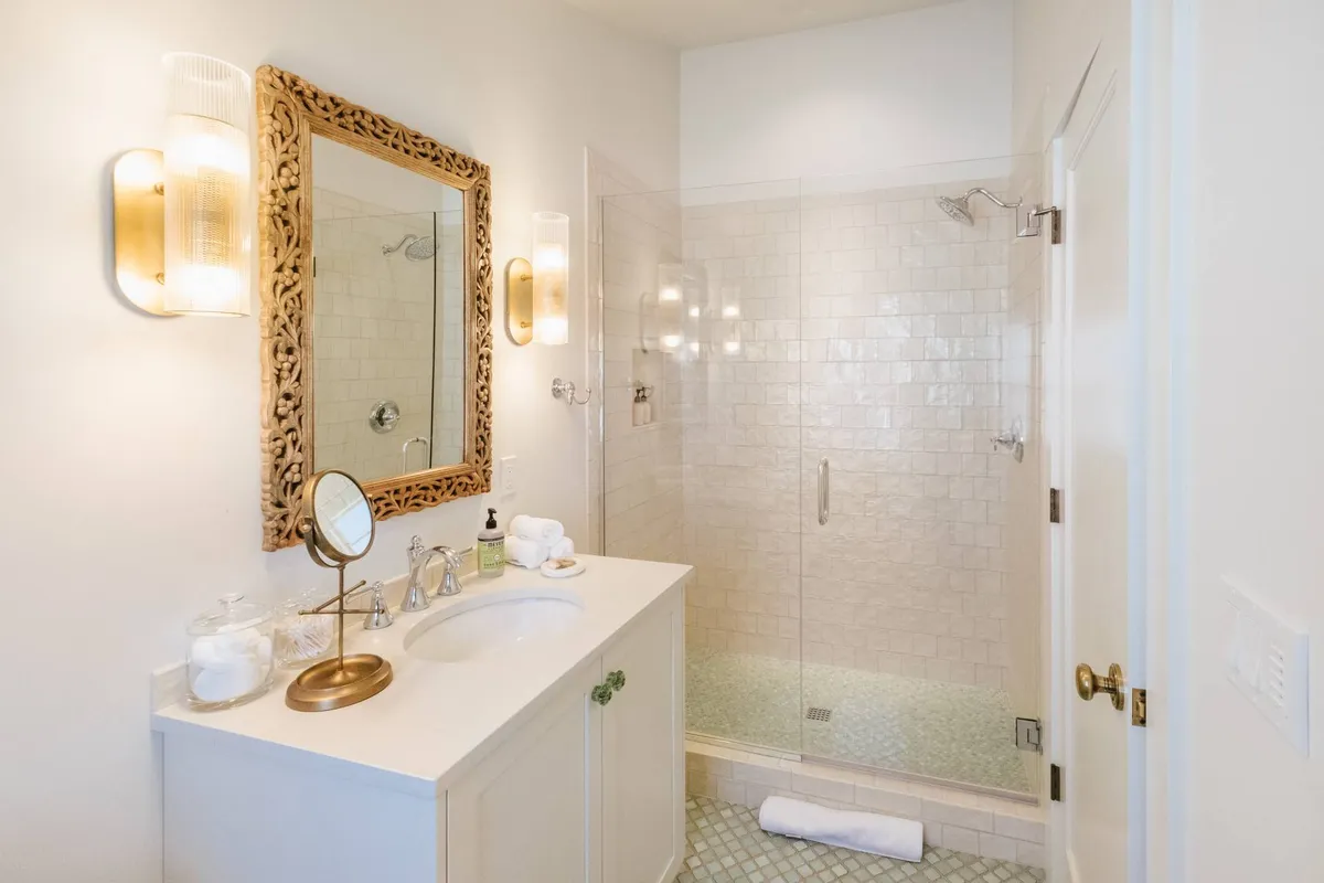 A calming bathroom scene with an ornate mirror, glass shower enclosure, and a neat vanity with a sink. Morning sunlight streams in, highlighting the clean, spa-like environment and suggesting a space designed for rejuvenation and self-care.