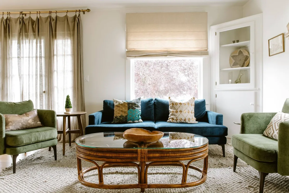 The image showcases a stylish, sunlit living space with a plush blue sofa, inviting green accent chairs, and a round glass coffee table set atop warm hardwood floors. The room feels both modern and cozy, blending fresh decor with touches of classic Craftsman detail. Natural light streams in, creating a bright, welcoming environment perfect for relaxation or socializing.