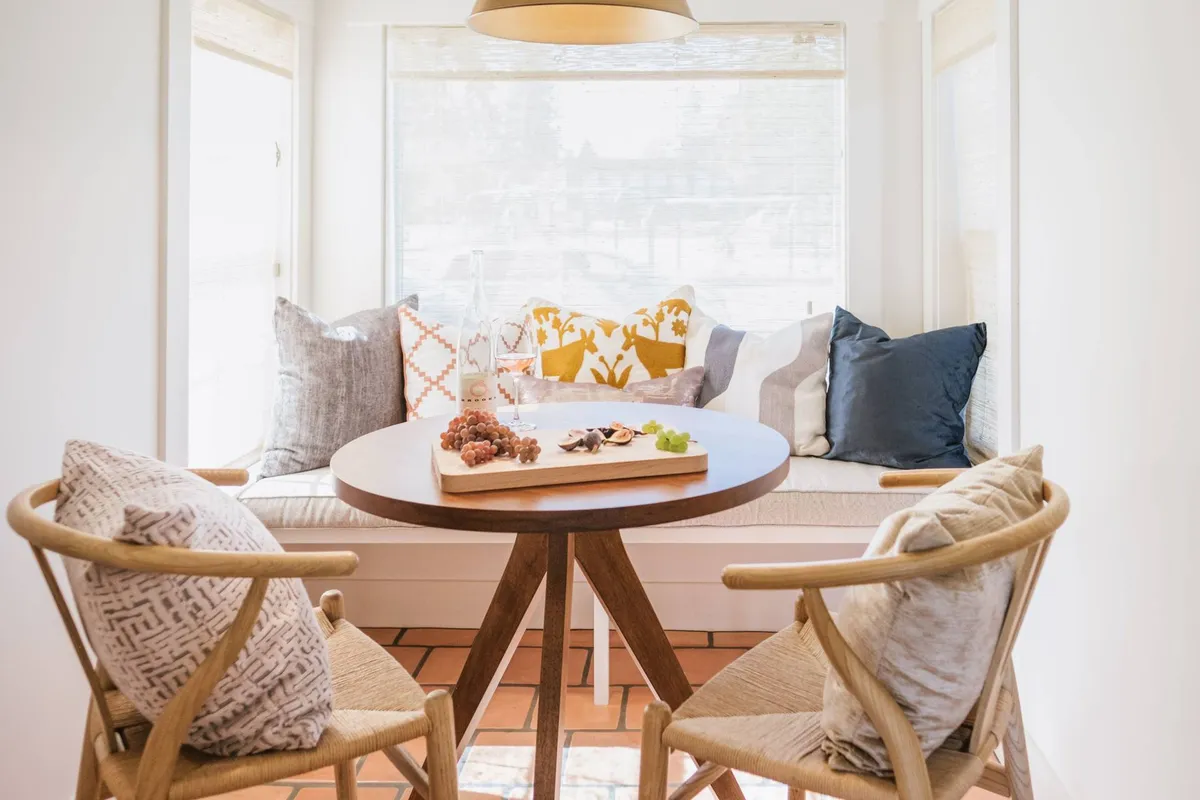 A bright and inviting dining nook featuring a round table surrounded by wooden chairs and a cushioned bench with decorative pillows. A tray with snacks sits atop the table, and large windows let in ample natural light, offering a glimpse of the outdoors and suggesting a pleasant view—possibly of Rotary Park.