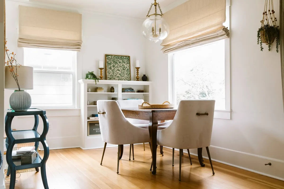 This image features a warm and inviting dining area with a comfortable table and chairs, decorative wall art, and a bookshelf filled with decor. The space is filled with natural light, creating a communal and welcoming atmosphere ideal for gathering and planning outings.