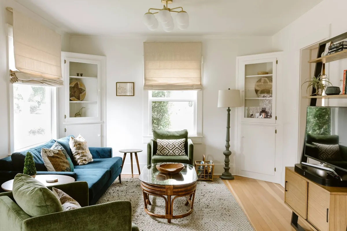 A spacious, comfortable living room featuring a blue sofa and green armchair arranged around a glass coffee table. Decorative pillows add a touch of warmth, while the layout is open and inviting—perfect for relaxing, entertaining, or gathering with friends. Large picture windows (not directly visible but suggested by the brightness and setup) flood the space with natural light, creating a luminous atmosphere.