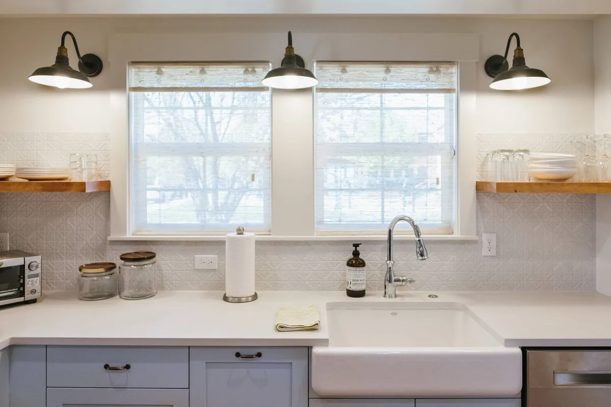 A bright, functional kitchen with ample windows, shelving, and lighting fixtures. The scene highlights the sink area with open shelves and a cozy, communal vibe—perfect for preparing meals or snacks before a family day out.
