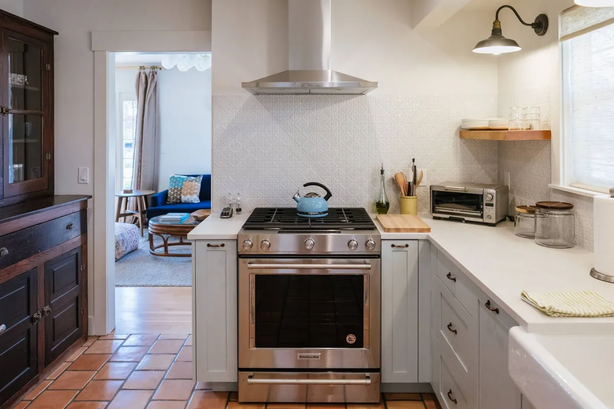 A stylish, well-equipped kitchen with a stainless steel stove, blue kettle, wooden countertop, and glass jars. The space feels cozy, with touches that blend Craftsman charm and eclectic high-end flair, and is clearly designed for socializing and gourmet cooking.