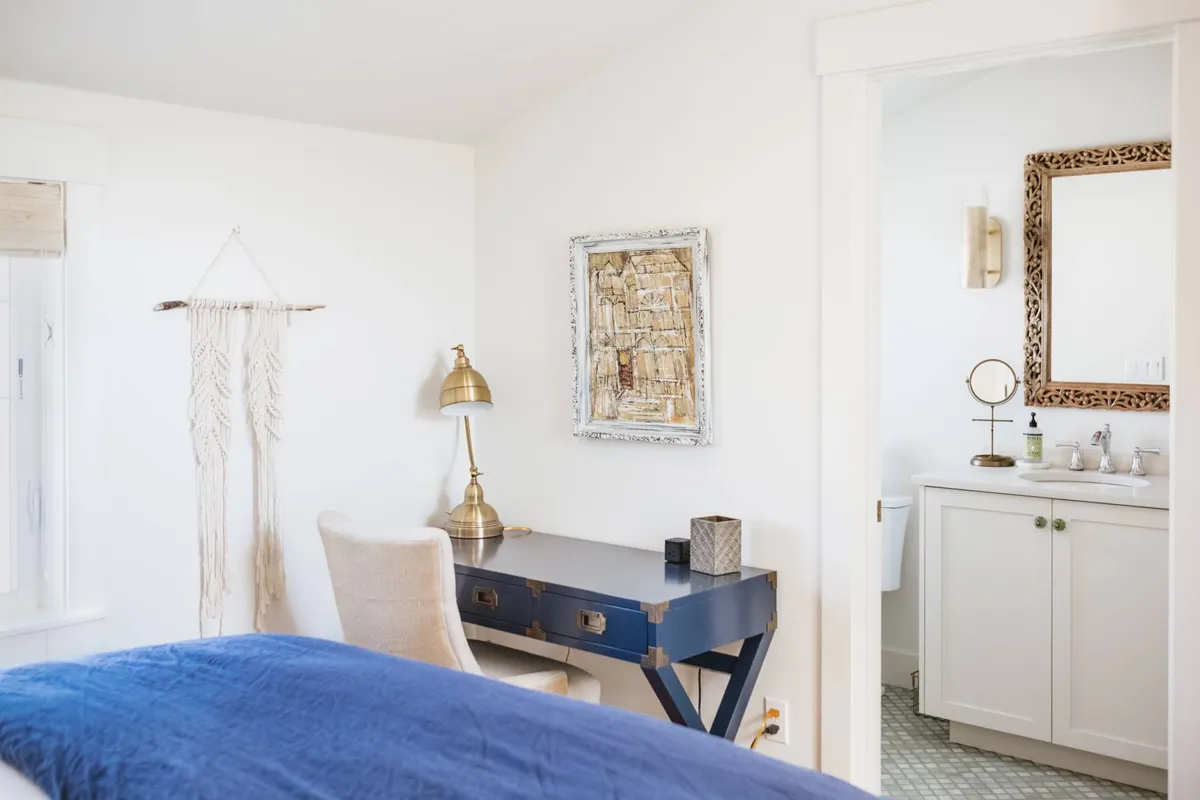 This image features a serene bedroom with a blue bedspread, a work desk with a lamp, framed artwork, and a visible entrance to the bathroom. The room is tidy and softly lit, with inviting linens and a tranquil color palette that communicates both comfort and functionality. The visible bathroom entrance subtly reinforces the en-suite feature.