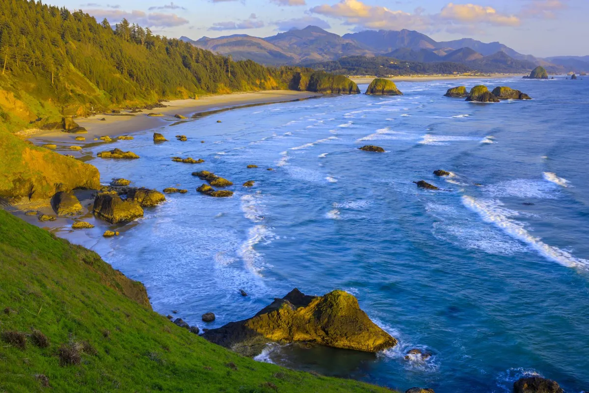 A tranquil exterior shot capturing a dramatic rocky coastline, gentle waves, distant forested mountains, and a wide expanse of sandy beach. The natural beauty of the setting invites exploration and relaxation, evoking a sense of adventure and connection to the outdoors.